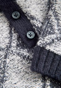 Jersey de punto en gris y blanco con un patrón texturizado. Cuenta con cuatro botones azul oscuro y puños acanalados en azul oscuro.