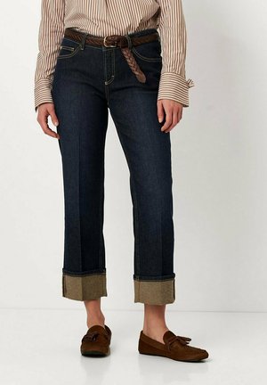 Dark denim jeans with a straight cut, featuring a braided brown belt and rolled hem with contrasting tan fabric. Brown moccasin shoes.