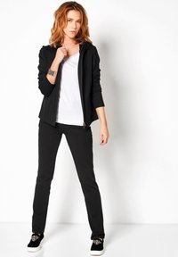 Zwarte zip-up jack, witte t-shirt, zwarte broek en versierde zwarte sneakers. Glad materiaal, getailleerd ontwerp en enkellange broeken.