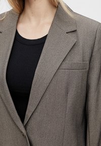 Blazer gris texturé avec revers crantés et une poche poitrine, superposé sur un haut noir côtelé. Fermé par un bouton.