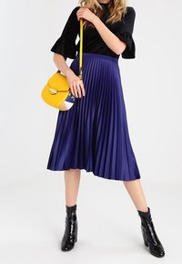 Gonna plissettata blu royal con una texture liscia, abbinata a una maglietta nera, una borsa gialla e stivali neri con tacco.