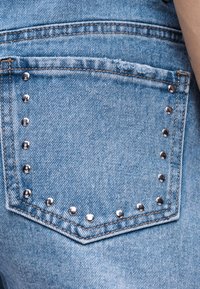 Τσέπη από μπλε τζιν με ασημένια μεταλλικά τρουκς που περιγράφουν την άκρη της τσέπης και μια φθαρμένη υφασμάτινη λεπτομέρεια κοντά στη ραφή στο πάνω μέρος.