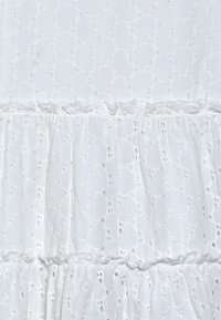 Biały bawełniany materiał z haftem ażurowym, z wzorami w kształcie okręgów i falbaną na dole. Tekstura jest lekka i przewiewna.