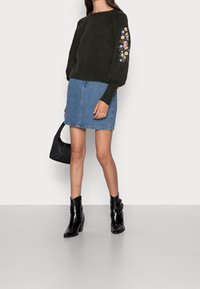 Pull en tricot vert avec broderies florales sur les manches, associé à une jupe en denim, des bottines noires à talons, et un petit sac à main noir.