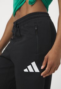 Melnas sporta bikses no mīksta materiāla ar elastīgu jostu, sānu rāvējslēdzēja kabatām un baltu Adidas logo uz augšstilba.