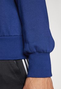 Blå sweatshirt med en texturerad tyg, som har en ihopdragen mudd. Designen inkluderar en ribbad nederdel och är kombinerad med svarta byxor med ränder.
