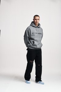 Gray hoodie with a kangaroo pocket and black logo text. Paired with black ripped jeans and blue and white sneakers.