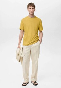 Gul kortærmet t-shirt parret med lysebeige linnedbukser. Manden holder en matchende blazer og har sorte sandaler på. Simpelt design.