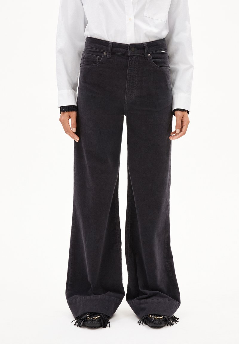 Femme portant un pantalon noir taille haute à jambes larges, des chaussures noires à franges et une chemise blanche à manches longues boutonnée.