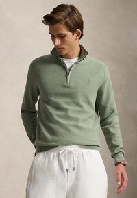 Joven con un suéter verde claro con cremallera hasta el cuarto, sobre una camisa blanca y pantalones blancos con cordón en la cintura, mirando hacia abajo con las manos en los bolsillos.