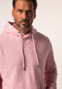 Sudadera rosa con un bolsillo frontal, cordones y un pequeño logo gris en el pecho. La tela parece suave y ligeramente texturizada.