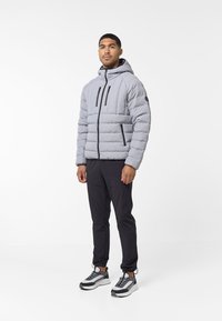 Giacca a vento grigia con texture a righe verticali, cerniera frontale, due tasche nere con zip, indossata sopra pantaloni sportivi neri e sneakers grigie.