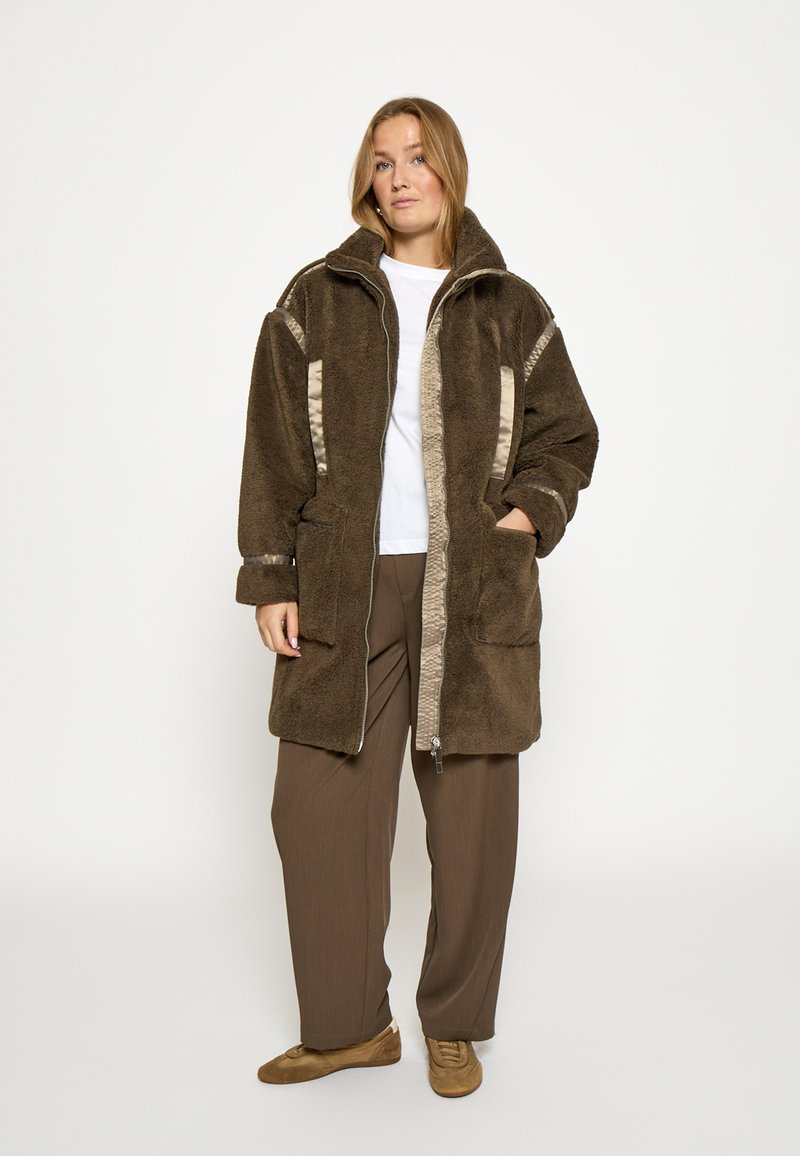 Hnedá fleecová bunda s vysokým golierom, kontrastnými béžovými detailmi, zapínaním na zips a bočnými vreckami, skombinovaná s voľnými hnedými nohavicami.