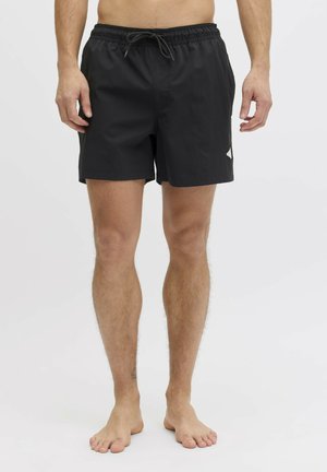 Homme portant un short de bain noir à cordon, debout pieds nus devant un fond blanc uni.