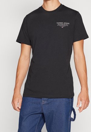 Czarny t-shirt Tommy Jeans z krótkim rękawem i białym logo na lewej piersi, noszony z niebieskimi dżinsami.