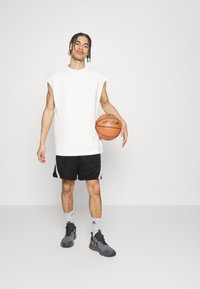 Weißes ärmelloses Sportoberteil, schwarze Basketballshorts mit seitlichen Akzenten, graue Turnschuhe, hält einen Basketball in der Hand, steht vor einem schlichten Hintergrund.