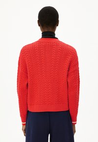 Persoon in een felrode, textuurige kabelgebreide trui over een zwarte turtle neck, gecombineerd met donkerblauwe broek, gezien vanaf de achterkant.
