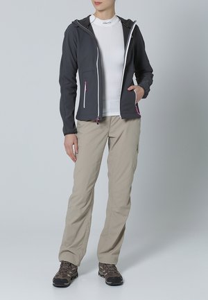 Giacca a cappuccio grigia con cerniere bianche e dettagli rosa, indossata sopra un top bianco aderente e pantaloni cargo beige. Stivali da trekking marroni completano il look.
