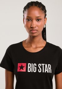 Tânără cu părul împletit poartă un tricou negru cu o stea roșie și text alb „BIG STAR” pe față, fundal neutru.