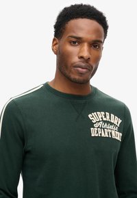 Grøn sweatshirt med cremefarvet tekst "Superdry Athletic Department". Har hvide sidestriber og en rund halsudskæring. Blød stofstruktur.