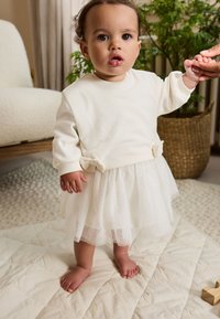 Weißes Langarmoberteil mit weicher Textur, kombiniert mit einem durchsichtigen, glitzernden weißen Tüllrock. Das Kind steht barfuß auf einer strukturierten, hellen Oberfläche.