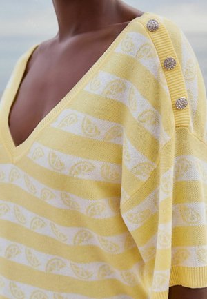 Sweter w serek w żółto-białe paski z wzorami plasterków cytryny i trzema ozdobnymi guzikami na ramieniu.