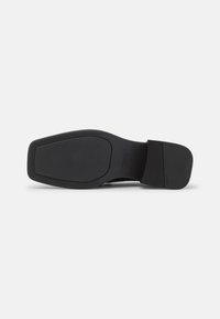 Semelle de chaussure noire à bout carré avec des zones de grip texturées au talon et à l'avant, présentée sur un fond clair uni.