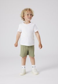 T-shirt bianco in cotone, pantaloni corti verde oliva e scarpe da ginnastica bianche con velcro, completati da calzini bianchi con dettagli neri.
