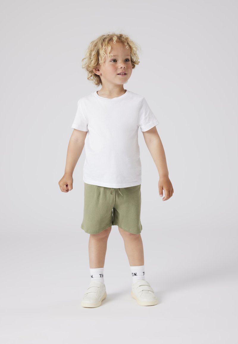 T-shirt bianco in cotone, pantaloni corti verde oliva e scarpe da ginnastica bianche con velcro, completati da calzini bianchi con dettagli neri.