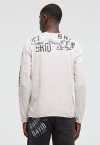 Helle beige Langarm-T-Shirt mit schwarzem grafischem Text und Designs auf dem Rücken, aus weichem Stoff. Mit Rundhalsausschnitt und gerippten Bündchen.