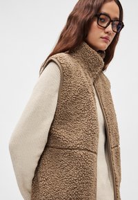 Καφέ γιλέκο από φλις με ψηλό γιακά και κλείσιμο με φερμουάρ, υφή υφάσματος, φορεμένο πάνω από εκρού πλεκτή μπλούζα. Ομαλές άκρες και διακριτικός σχεδιασμός.