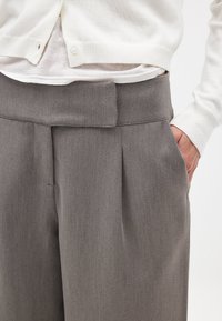 Pantalones gris con pliegues, de textura suave y un detalle en la cintura, combinados con un cárdigan blanco con acentos de botones.