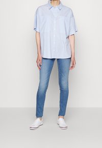 Camisa de botón de manga corta azul claro, con dos bolsillos frontales, combinada con unos pantalones vaqueros ajustados azul claro y zapatillas blancas. Diseño minimalista.