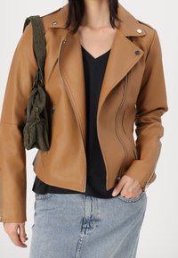 Hnědá syntetická kožená motorkářská bunda se zipovými kapsami, stříbrnými detaily a přiléhavým střihem, kombinovaná s černým topem s véčkovým výstřihem a džínovou sukní.