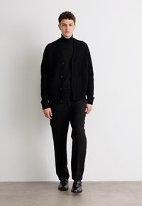 Μαύρο πλεκτό κασκόλ με ριγέ υφή, με κουμπιά μπροστά, φορεμένο πάνω από μαύρο λαιμόκοψη και μαύρο παντελόνι, συνδυασμένο με μαύρα παπούτσια.