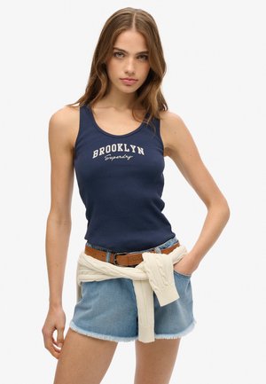 Débardeur navy avec texture côtelée et impression "BROOKLYN Superdry", associé à un short en denim clair et un pull crème noué à la taille.