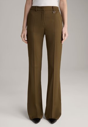 Femme portant un pantalon taille haute vert olive ajusté avec des plis devant, des passants de ceinture, et des chaussures noires à bout pointu, debout devant un fond uni.