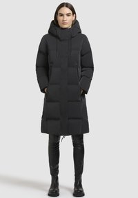 Zwarte pufferjas met een capuchon, knie-lange lengte, met een geborduurd ontwerp, voorzien van verticale naden en zijzakken. Gecombineerd met zwarte aangesloten broek en laarzen.
