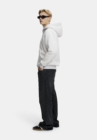 Lys grå hettegenser med en frontlomme, kombinert med svarte jeans. Modellen har på seg mørke solbriller og svarte sko, og står på siden.
