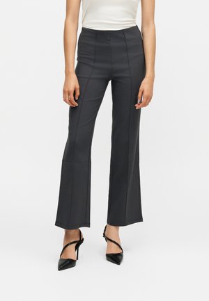 Femme portant un pantalon noir évasé taille haute, un haut blanc sans manches et des escarpins noirs pointus, debout devant un fond blanc uni.