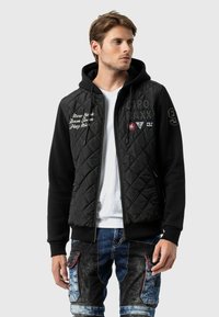 Schwarze gesteppte Jacke mit Reißverschluss und Kapuze, verziert mit weißen Buchstaben und Patches, kombiniert mit blauen Denim-Cargo-Hosen.