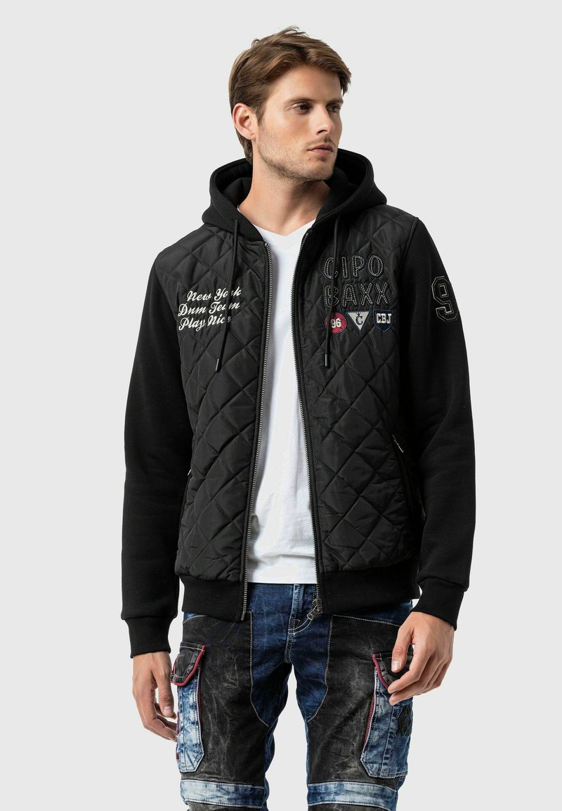 Zwart gewatteerd jack met een rits aan de voorkant en een capuchon, voorzien van witte letters en patches, in combinatie met blauwe denim cargo broek.