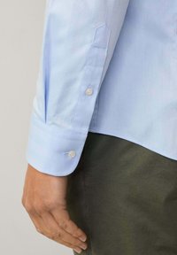 Camicia azzurro chiaro con texture a spina di pesce. Presenta polsini con bottoni, bottoni a contrasto e colletto a punta. Indossata con pantaloni verde scuro.