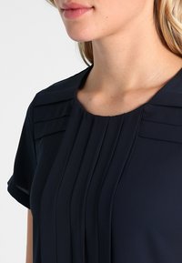 Námorní modrá blůza s krátkými rukávy, zdobená plizé kolem výstřihu, s jemnou texturou a zaobleným límcem.