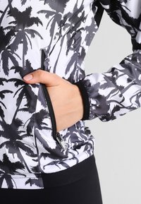 Chaqueta de cierre frontal en blanco y negro con un patrón de palmeras, un bolsillo en el frente, puños elásticos y una textura de tela lisa y brillante.