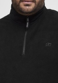 Svart fleece kvart-zip genser med høy krage, med en teksturert overflate, subtil logo-detalj og en nylon glidelås.