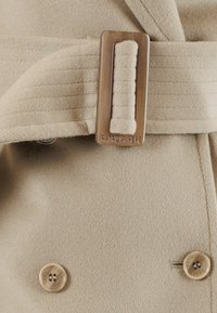Cappotto beige realizzato in materiale morbido con una cintura testurizzata dotata di una fibbia metallica rettangolare. Include un dettaglio a bottone sul lato.