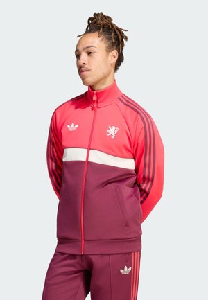 Mężczyzna z dredami ubrany w czerwono-bordową dresową kurtkę i spodnie Adidas, stoi ze złożonymi za plecami rękami, patrząc w lewo.