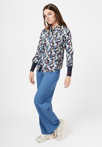 Blumenmuster-Hemd mit langen Ärmeln in Blau, Creme und Burgunder; kombiniert mit hellblauen Hosen und beigen Sneakers mit Netzdetail.