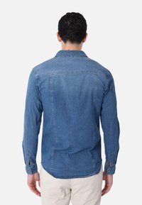 Camicia in denim azzurro chiaro, con maniche lunghe e polsini a bottoni, orlo curvo e dettaglio del giogo sul retro.